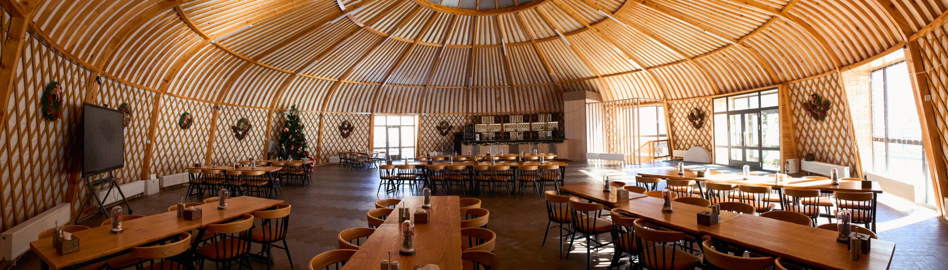 A spacious, circular room with a high, domed wooden ceiling. The room features long, wooden tables and chairs arranged neatly. Christmas wreaths adorn the walls, and a decorated tree stands in the corner. Large windows let in natural light.