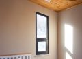 A minimalist bedroom with a wooden ceiling and a small window letting in natural light. The room has a bed with a gray blanket, white pillow, and a wooden side table. A geometric pendant light hangs from the ceiling.