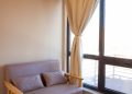 A cozy corner with a small beige sofa, two cushions, and large windows draped with beige curtains. The room features wooden flooring and a wooden ceiling, creating a warm and inviting atmosphere.