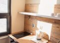 A small kitchen space with a wooden countertop, electric stovetop, and sink. There's a white kettle on a tray beside the sink. Sunlight streams in from a window on the left, illuminating the wood-paneled backsplash and a shelf with cups above.