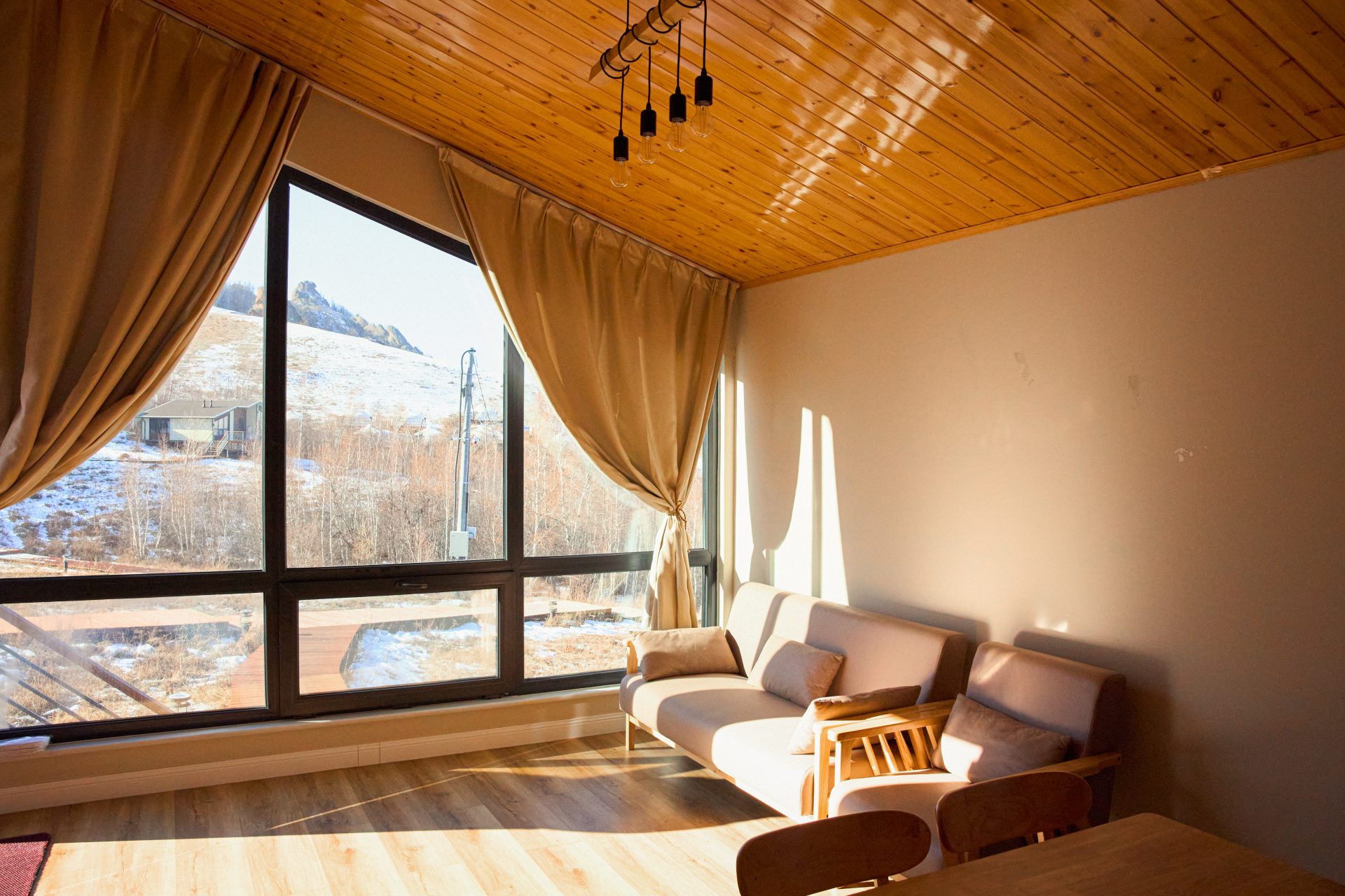 A cozy living room with a sloped wooden ceiling and large windows revealing a mountain view. Natural light floods the space, highlighting a beige sofa, wooden chair, and small table. Long curtains frame the scenery outside.