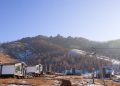 A wooden boardwalk leads uphill to modern cabins under a clear blue sky. The area is partially covered in snow, with a backdrop of rugged hills and sparse trees.