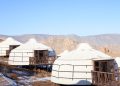 Three white yurts with wooden decks are situated on a snowy hillside, surrounded by dry grass and mountains in the background. A colorful cobblestone path leads toward the yurts under a clear blue sky.