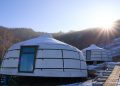 Two yurts in a snowy landscape with the sun shining brightly over a hill in the background. A pathway made of square tiles leads towards them, surrounded by snow. The scene is calm and picturesque, with clear blue skies.