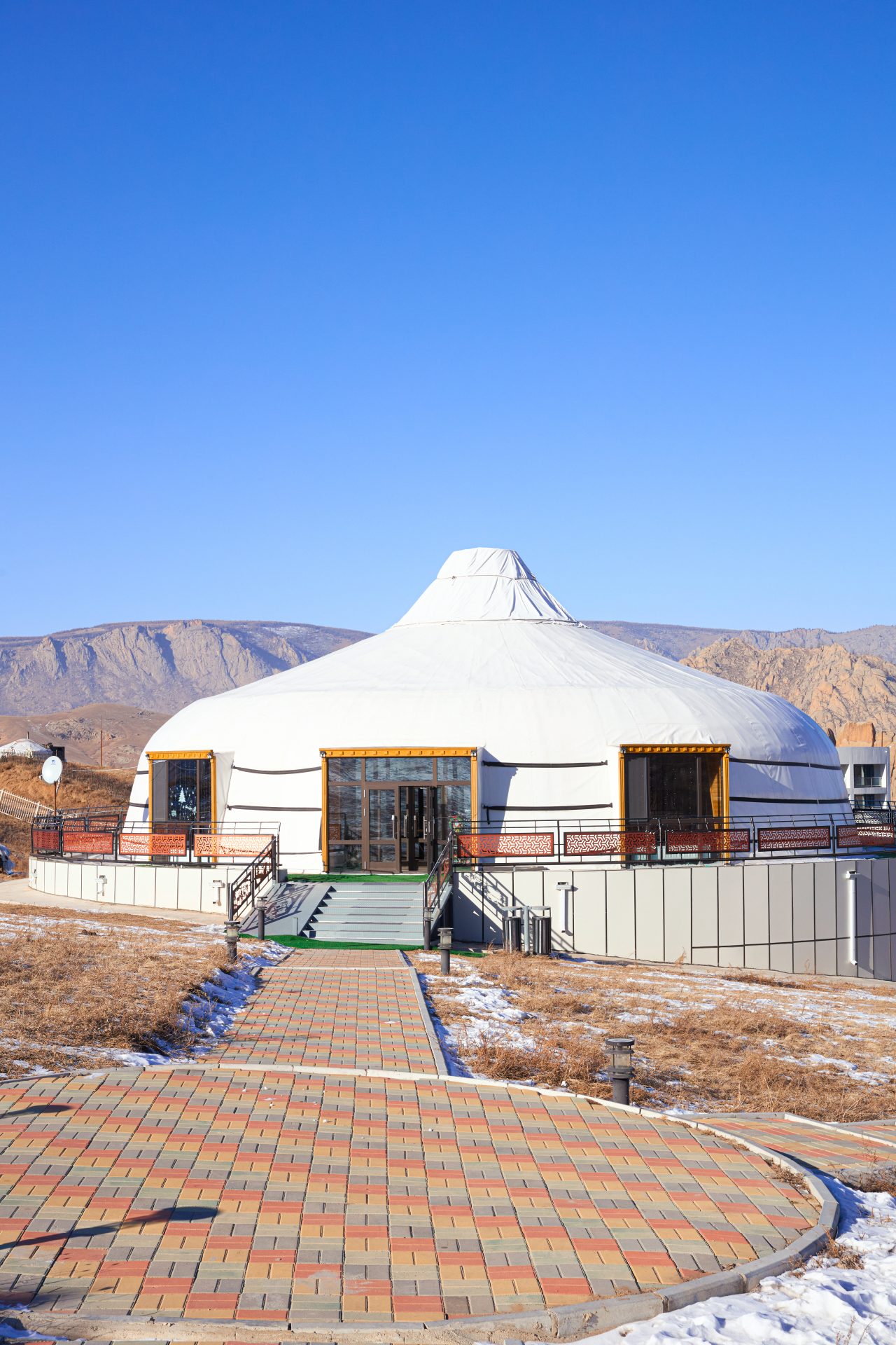 A traditional yurt with a white exterior and wooden windows sits under a clear blue sky. The structure is surrounded by dry grass and snow patches, with a paved pathway leading to its entrance. Mountains are visible in the background.