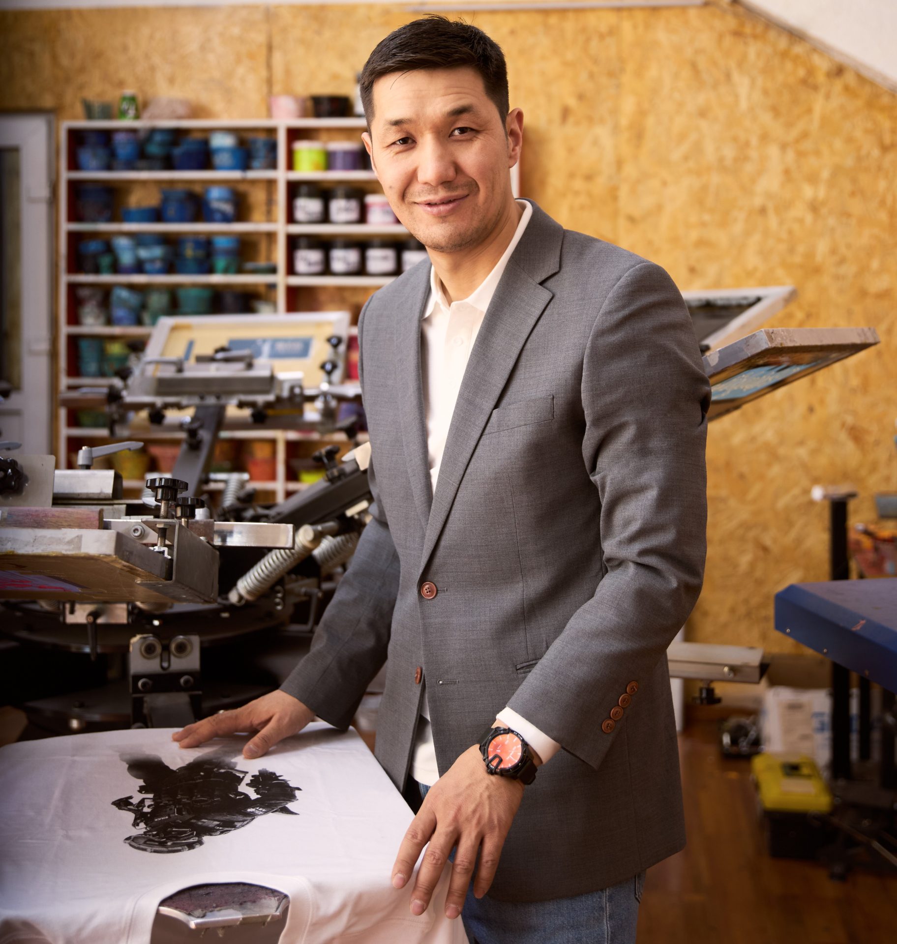 A man in a gray suit stands in a workshop, smiling at the camera. He is next to a screen printing machine with a white shirt on it. Shelves with colorful containers are visible in the background.