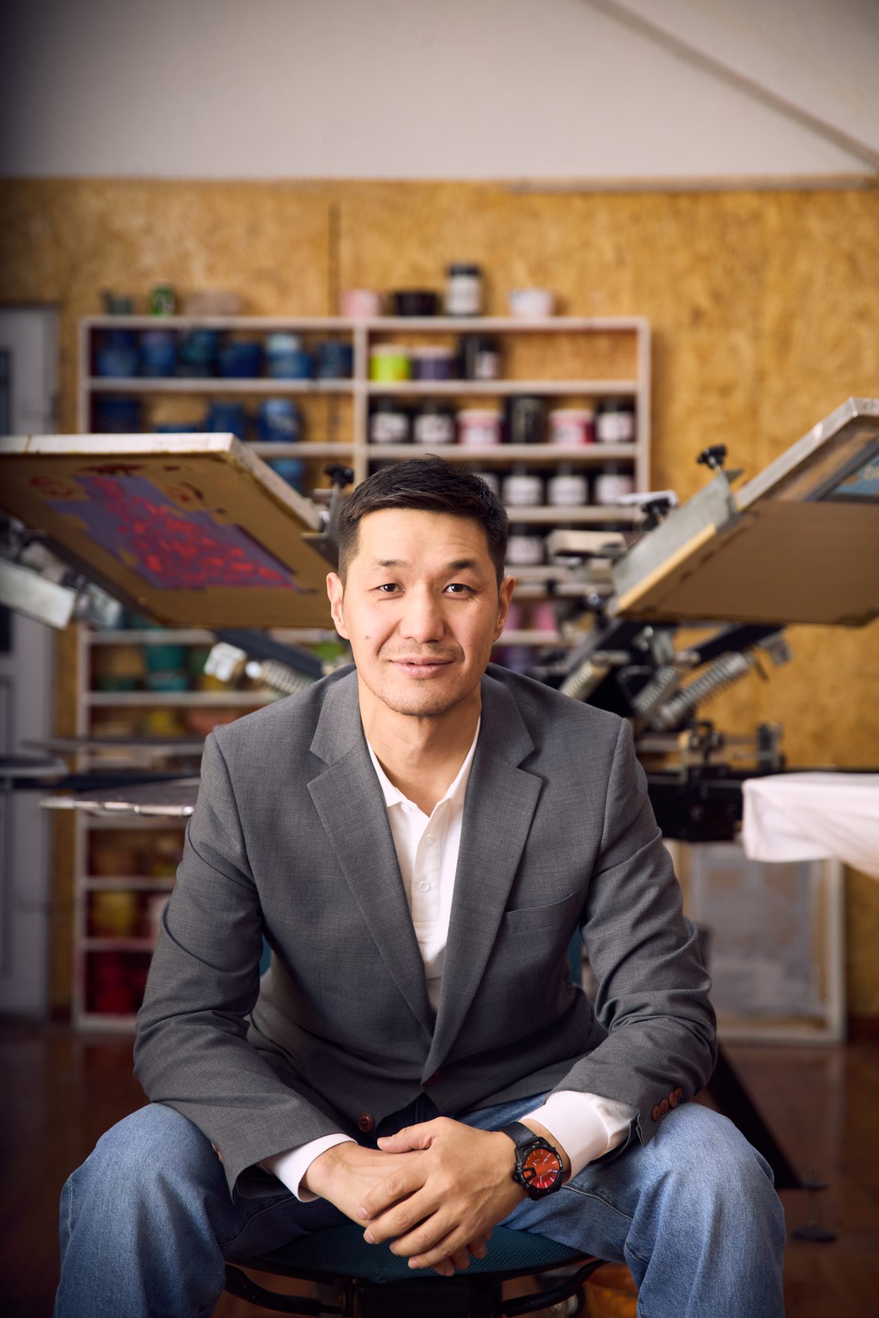 A person wearing a gray blazer and white shirt sits on a chair in a workshop. Behind them are shelves filled with paint cans and a screen printing press. The setting appears creative and industrious.