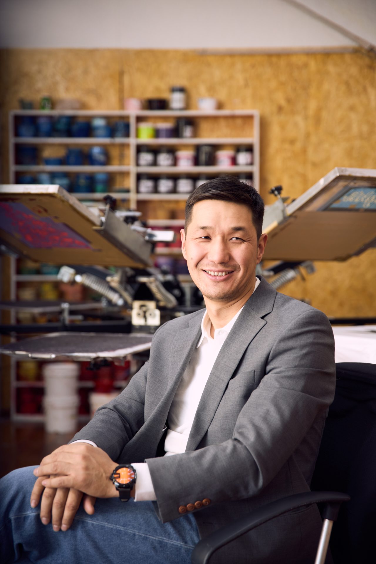 A person in a gray blazer and jeans sits smiling in front of a screen printing machine, with shelves of colorful supplies in the background.