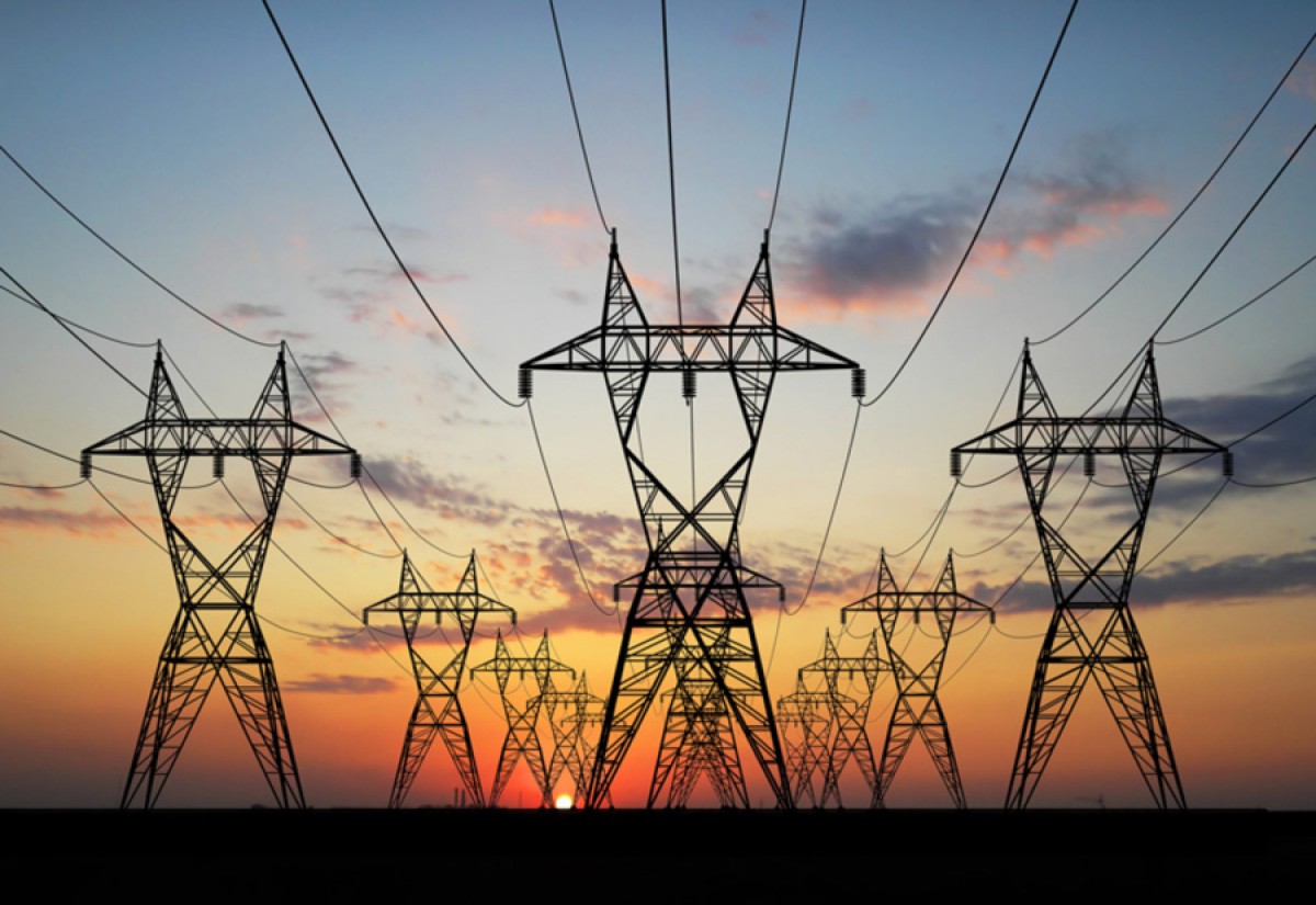 A row of electrical transmission towers silhouetted against a colorful sunset sky, with clouds scattered and the sun visible on the horizon. The towers and power lines create a striking pattern across the landscape.