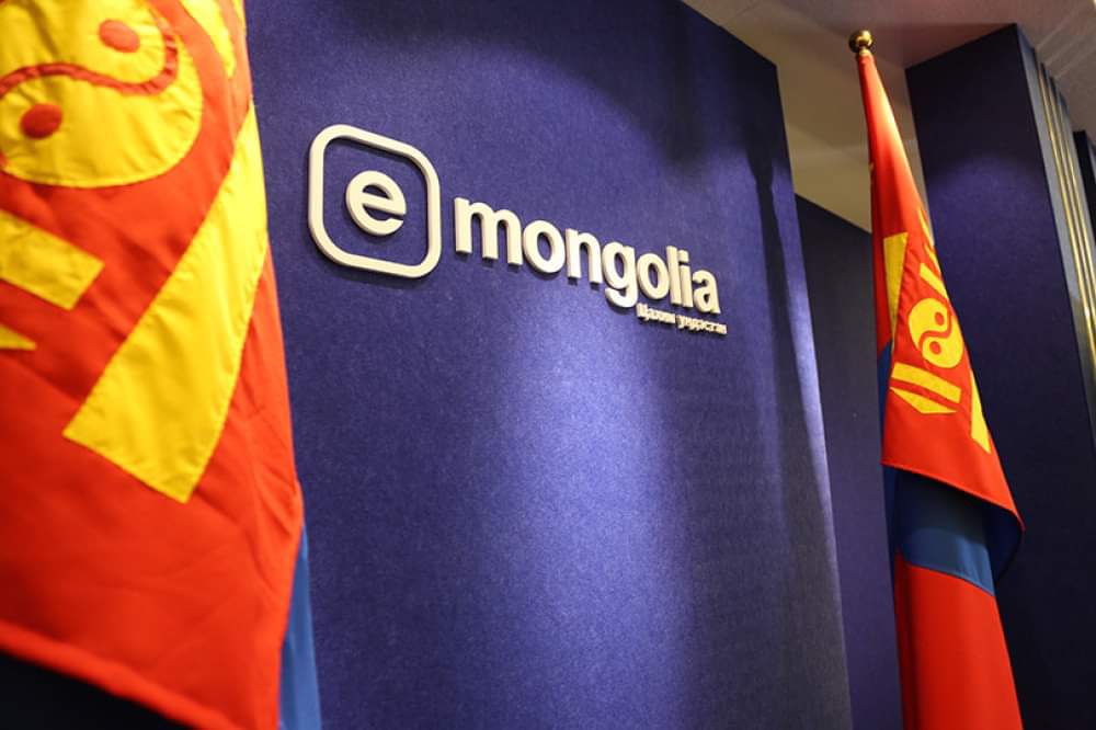 The image shows a blue wall with the logo and text "e-Mongolia" in white. Two Mongolian flags with red, yellow, and blue colors are visible on either side, partially framing the logo.