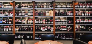 Shelves filled with a wide variety of wine bottles in a stylish wine shop. Black chairs and wooden tables are visible in the foreground, suggesting a tasting area. The arrangement is neat, and the lighting is warm and inviting.