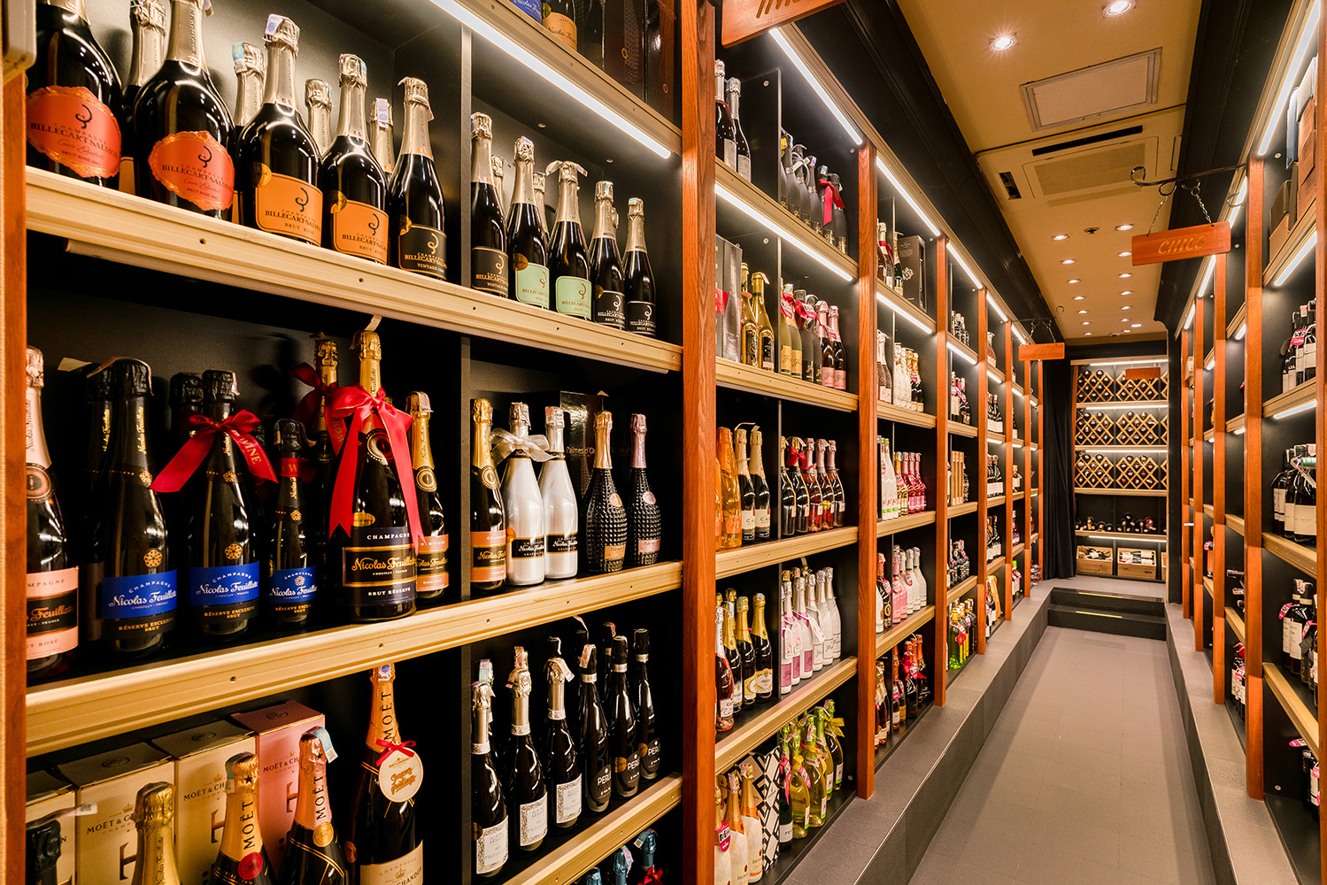 A well-lit wine cellar featuring neatly arranged bottles of champagne and wine on wooden shelves. The aisle leads to a wine rack at the far end, creating an organized and inviting atmosphere.