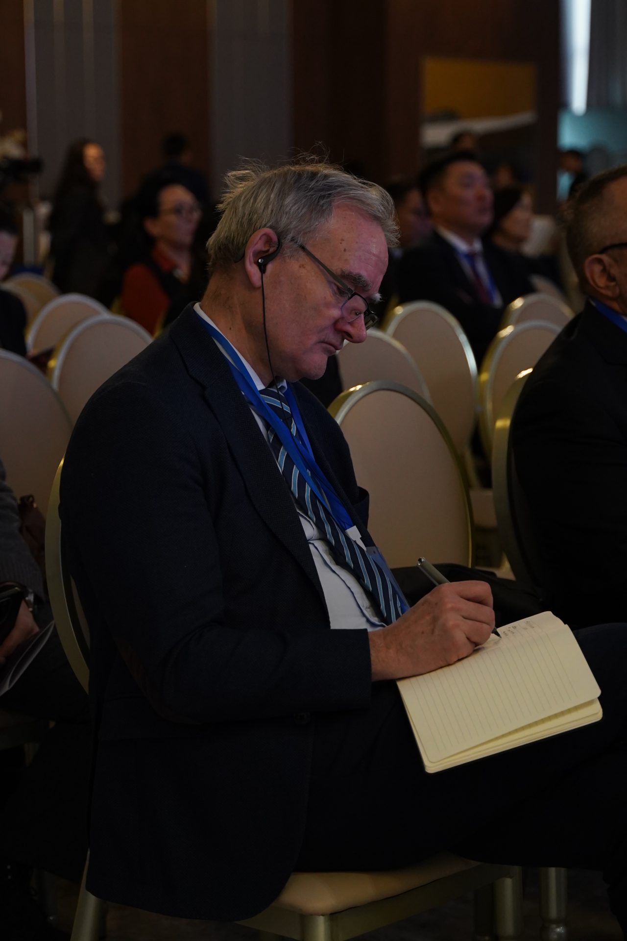 A man with glasses and headphones takes notes on a notepad while seated in a conference room. He wears a dark suit, striped tie, and a blue lanyard. The room is filled with other seated attendees.