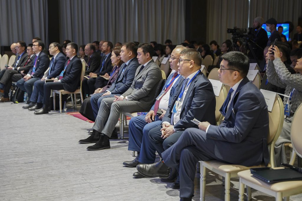 A group of people in business attire seated in rows at a conference, attentively facing the front. The room is well-lit, with beige curtains and carpet, and some attendees are taking notes or recording with devices.