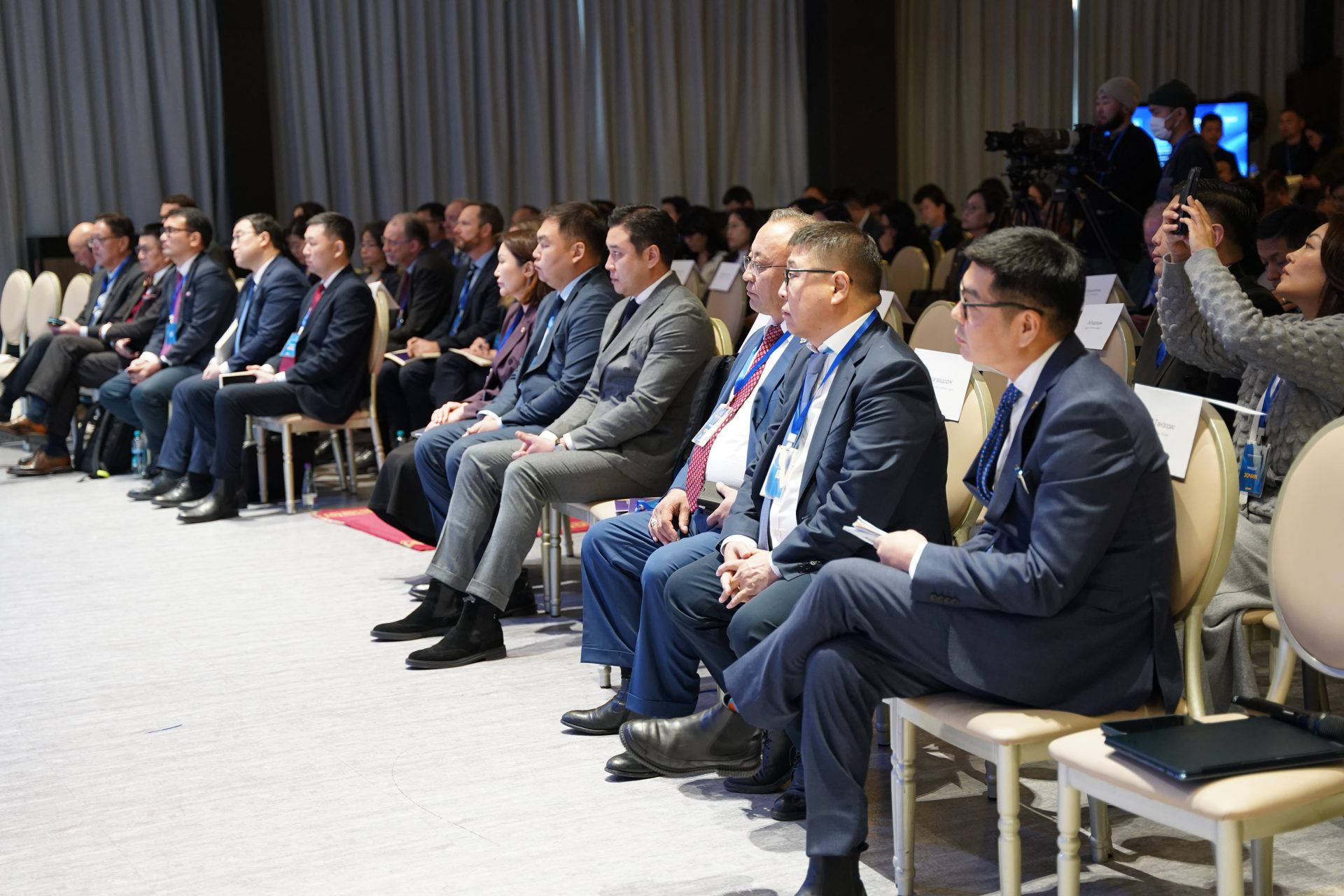 A group of people in formal attire sit attentively on chairs in a conference room. Attendees include men and women, some taking notes, with a camera setup in the background capturing the event.