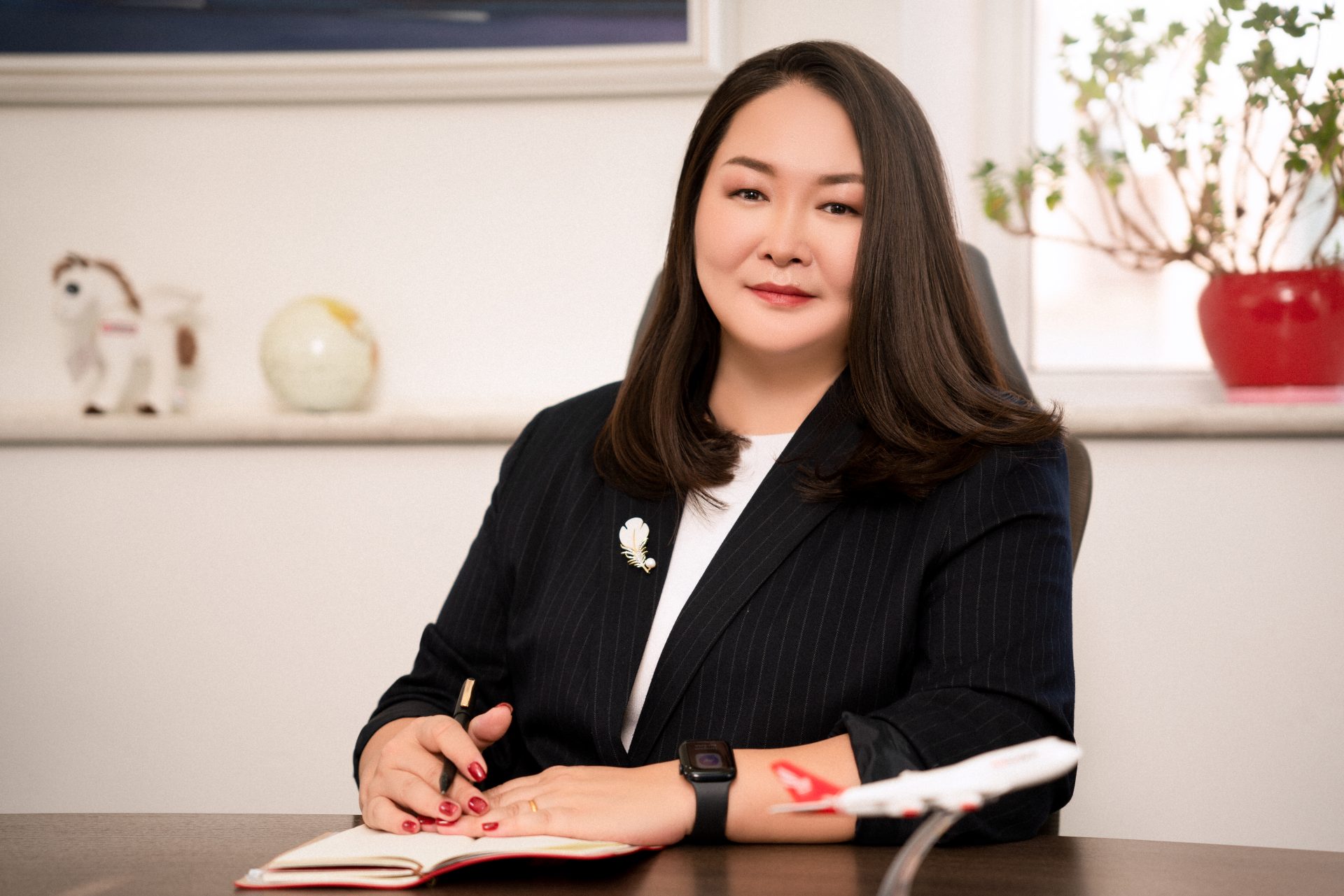 A woman with long dark hair sits at a desk, wearing a black pinstripe blazer and a brooch. She's writing in a notebook with a pen. A toy airplane, globe, and plush horse are in the background on a white shelf.