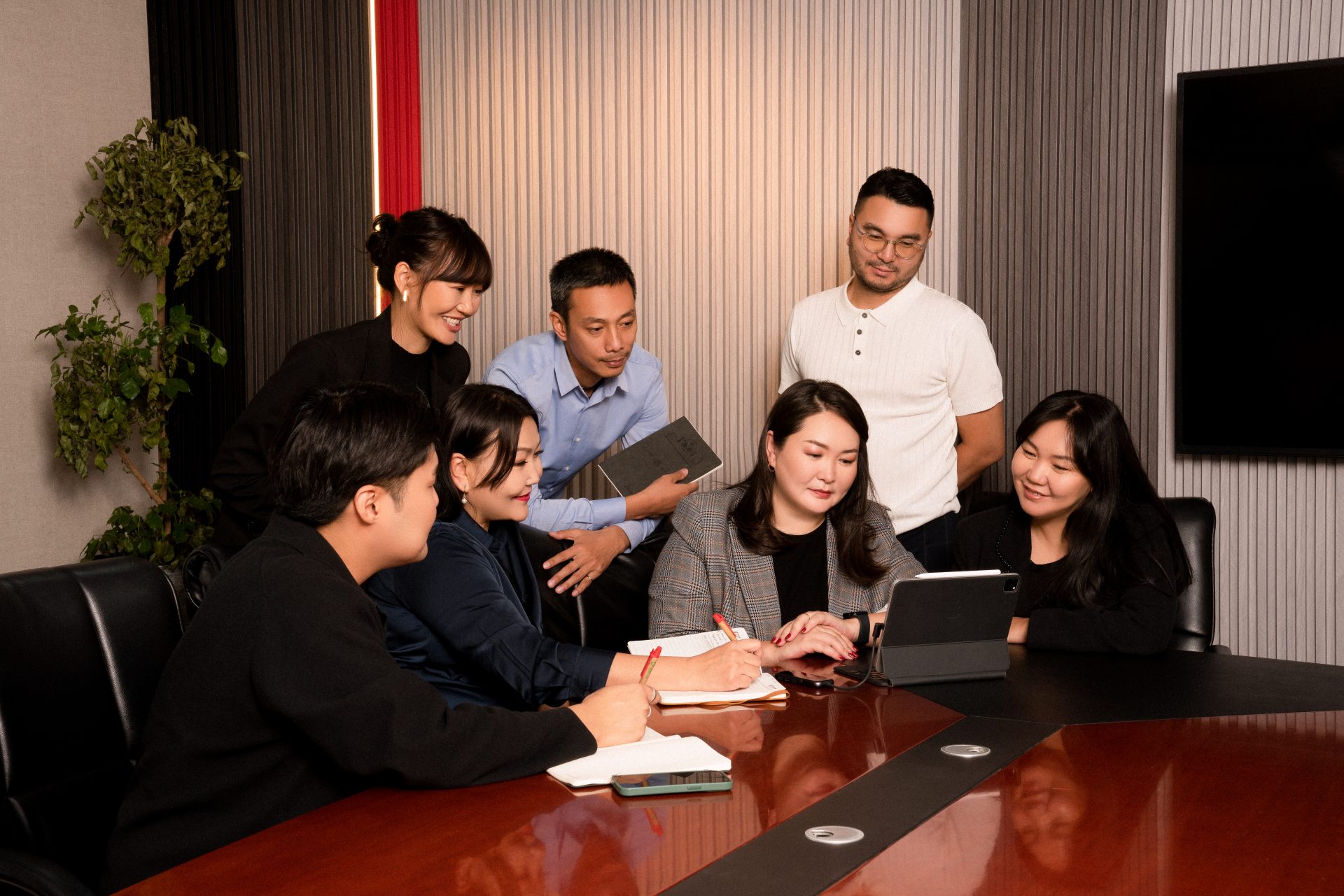 A group of seven people sitting and standing around a table in a conference room. They are looking at a tablet, engaging in discussion. Notebooks and pens are visible on the table. The room has a modern design with a large screen on the wall.