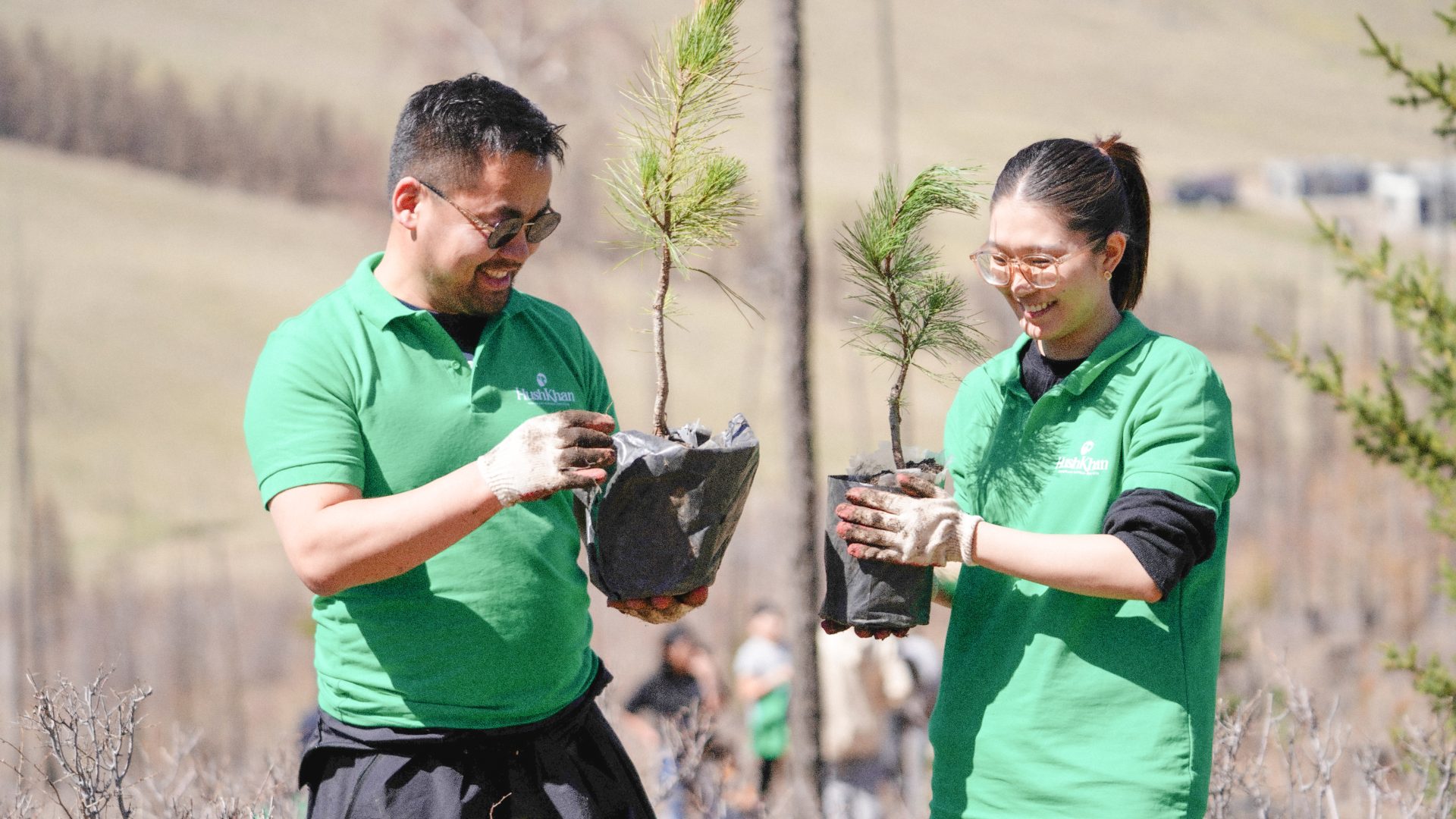 Two people in green shirts and gloves hold small potted trees. They are smiling and standing in an outdoor setting, possibly participating in a tree planting event.