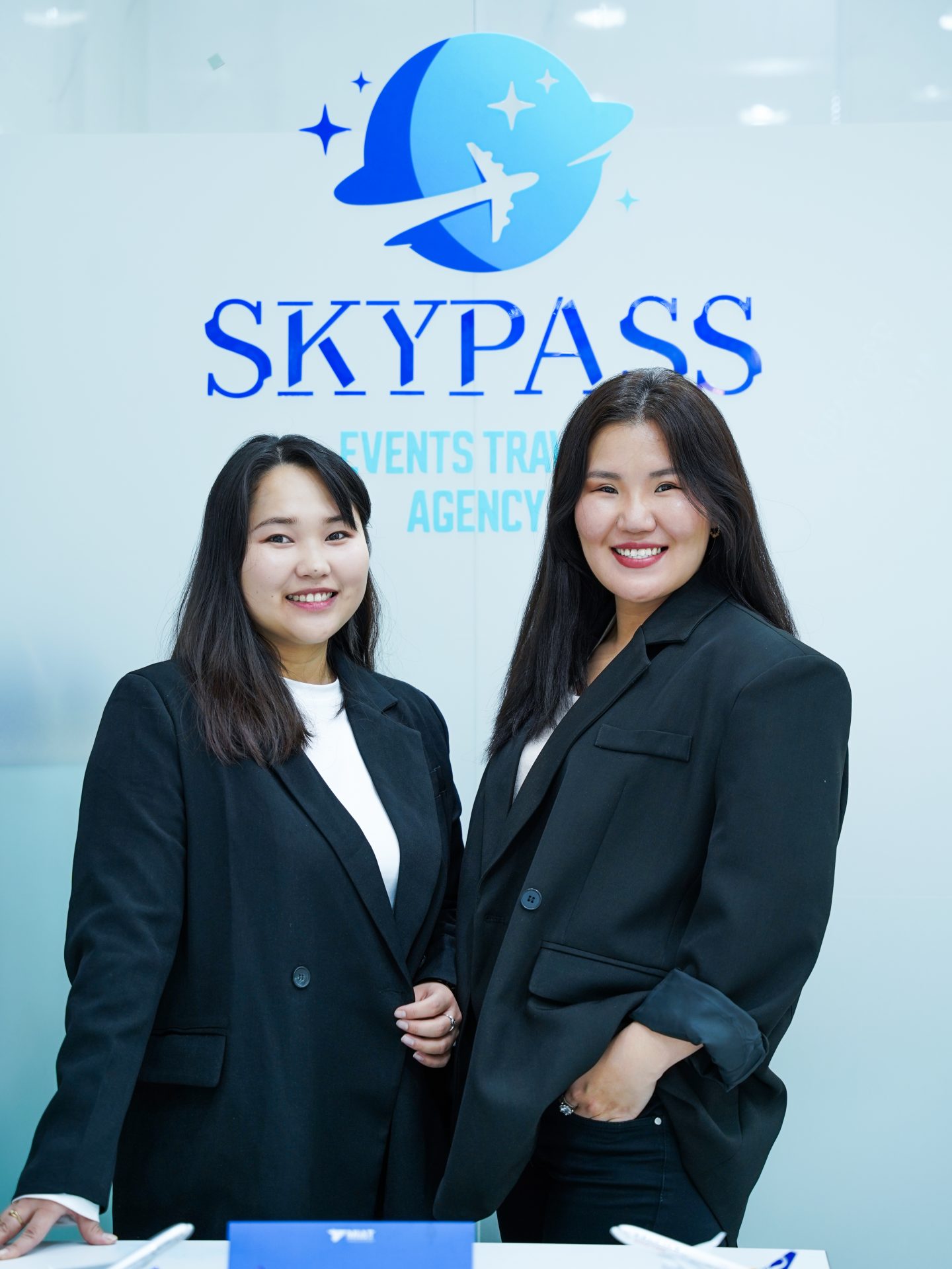 Two women in black blazers stand smiling in front of a "Skypass Events Travel Agency" sign. The woman on the left has long, straight hair, while the woman on the right has her hand in her blazer pocket.