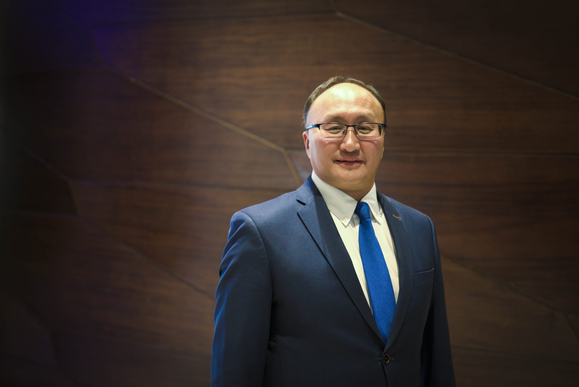 A man in a blue suit and tie stands in front of a wooden paneled wall. He is wearing glasses and has a neutral expression.