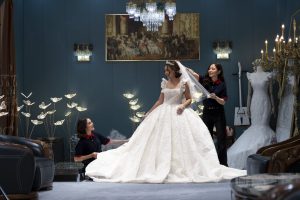 A bride in a white gown stands in a luxurious room with elegant decor. Her Онцгой мөчийн хамтрагч attendants adjust her veil, surrounded by chandeliers and mannequins in wedding dresses. The setting is opulent and serene.