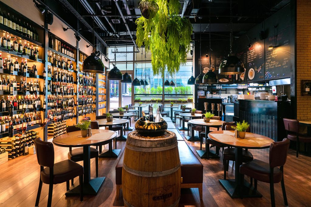 A cozy restaurant interior showcases wooden tables and chairs around a wine barrel centerpiece, embodying дарсны соёл бол хүн төрөлхтний үнэт өв. Shelves brimming with wine bottles line the wall, while a lush hanging plant adds greenery and large windows flood the space with natural light.