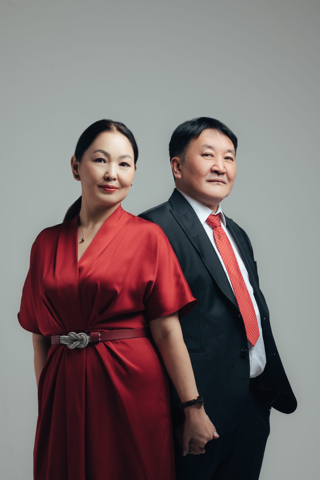 A couple stands back-to-back against a neutral background. The woman wears a red dress with a decorative belt, and the man is in a dark suit with a red tie. They both have a confident and relaxed expressions.