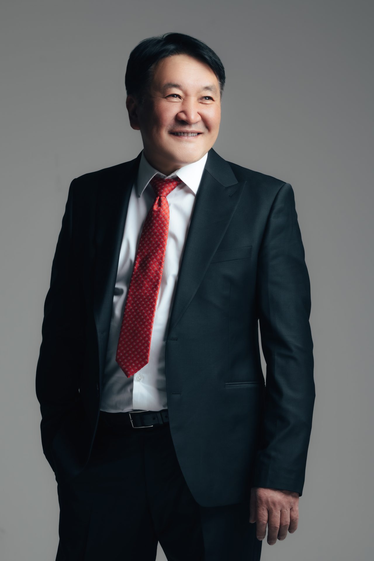 A man in a dark suit and red tie smiles, standing with one hand in his pocket. The background is a neutral gray.