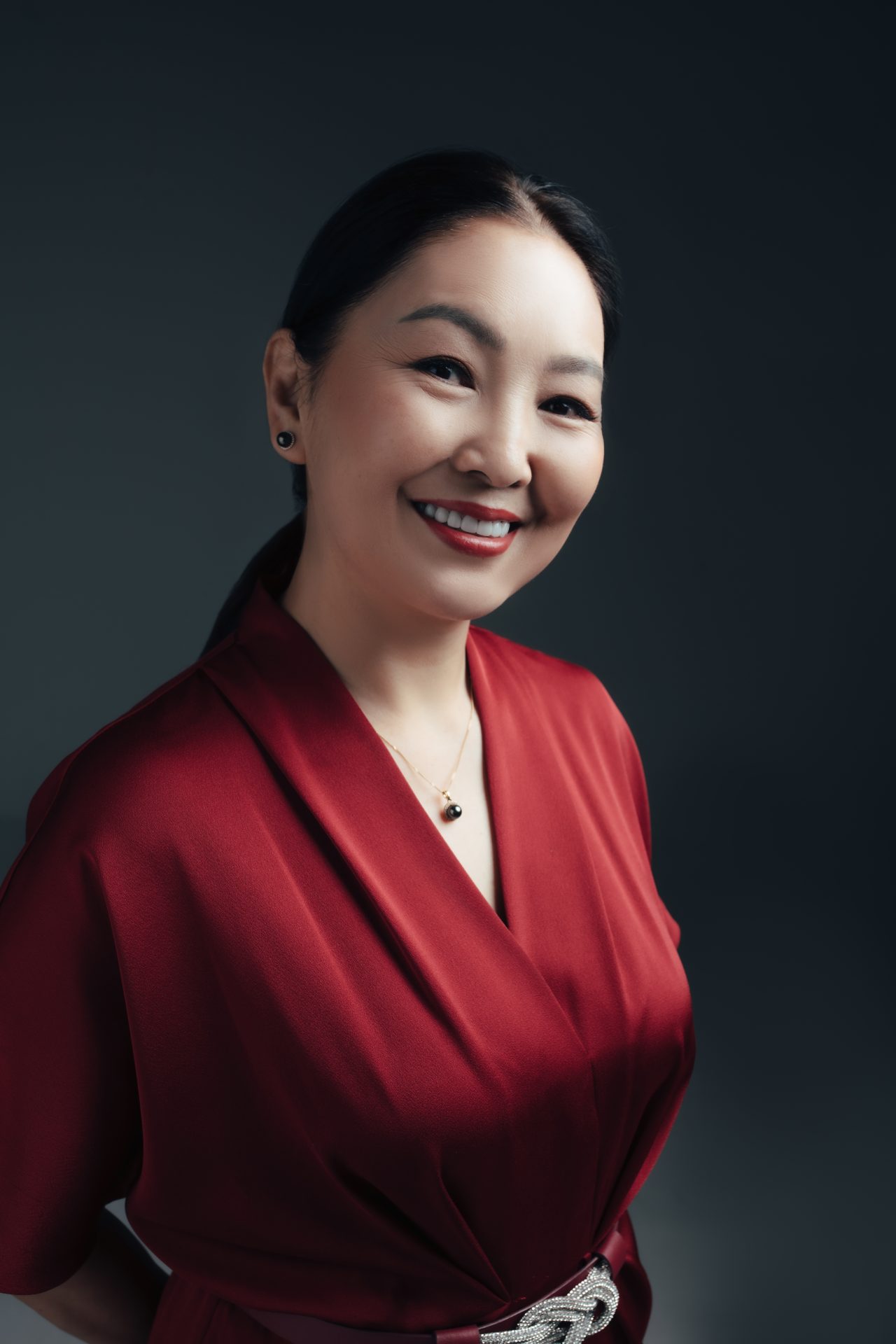 A smiling woman wearing a red outfit stands against a dark background. She has long, dark hair and wears a necklace and earrings. Her hands are positioned behind her back.