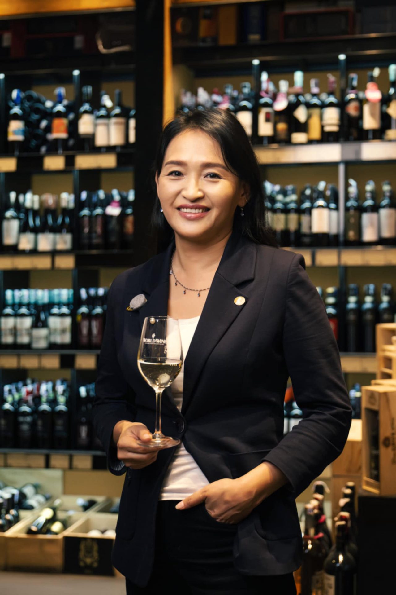 A woman in a formal outfit stands in a wine shop, smiling and holding a wine glass, embodying "дарсны соёл бол хүн төрөлхтний үнэт өв." Shelves brimming with wine bottles form the backdrop of this rich tapestry of human heritage.