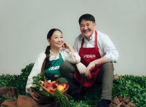 A woman and a man, both wearing aprons with "VIDAN" on them, are smiling while surrounded by lush green foliage. The woman holds a basket of vegetables, including carrots and bell peppers. They both appear happy and content.