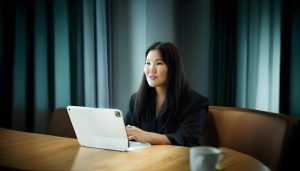 A woman sits at a wooden table using a tablet with a keyboard. She is wearing a dark blazer and is smiling. A cup is placed on the table in front of her. The background has dark curtains and a dim ambiance.