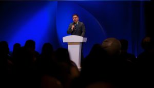 A man in a suit stands at a white podium, speaking into a microphone against a blue-lit backdrop. The audience is in shadow, creating a contrast with the brightly lit stage.