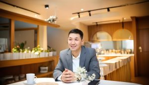 A man in a suit sits at a table in a modern, warmly lit cafe. He is smiling and speaking, with a cup of coffee and a smartphone on the table in front of him. The background shows decorative plants and interior lights.