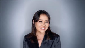 A woman with dark hair smiles at the camera. She is wearing a black blazer and has a neutral-colored background.