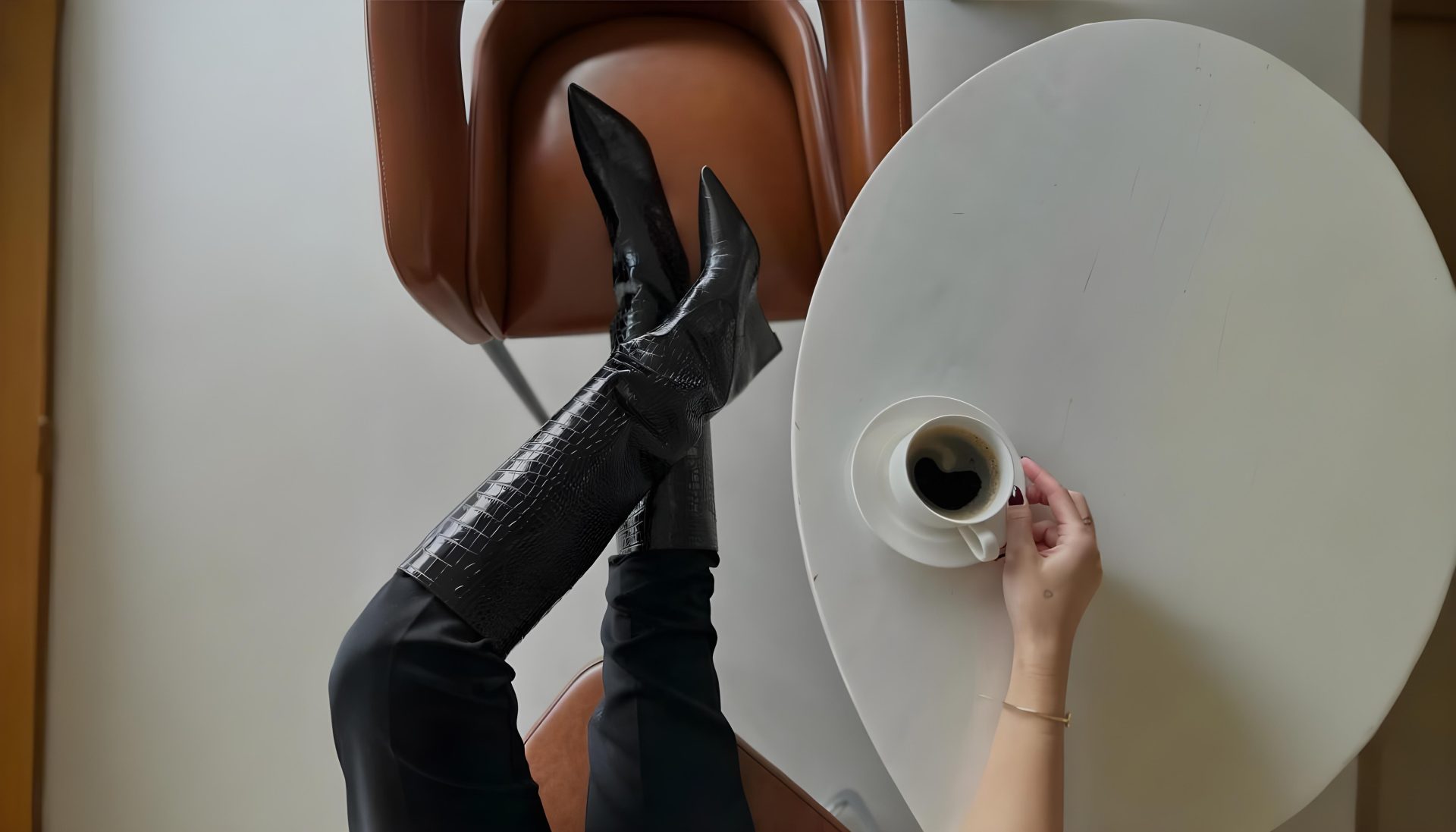 A person is lounging with their legs up on a chair, wearing black leather boots. They are holding a cup of coffee over a round white table. The setting is indoors with a minimalist design.