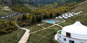 A scenic view of Sulo Resort featuring a group of white yurts nestled in a lush, hilly landscape. A large yurt with a deck is in the foreground, next to a fenced tennis court. Several cabins are scattered among the trees.