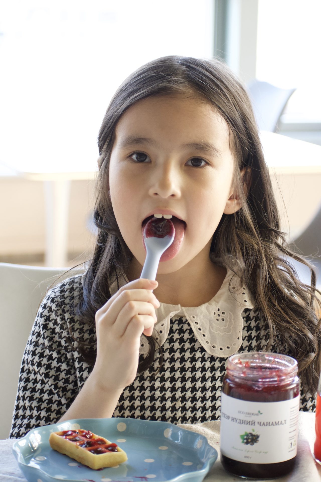 A young girl with long dark hair is eating jam with a spoon. She is sitting at a table with a blue plate in front of her, which holds a slice of bread topped with jam. An open jar of jam sits nearby. The background is softly lit.