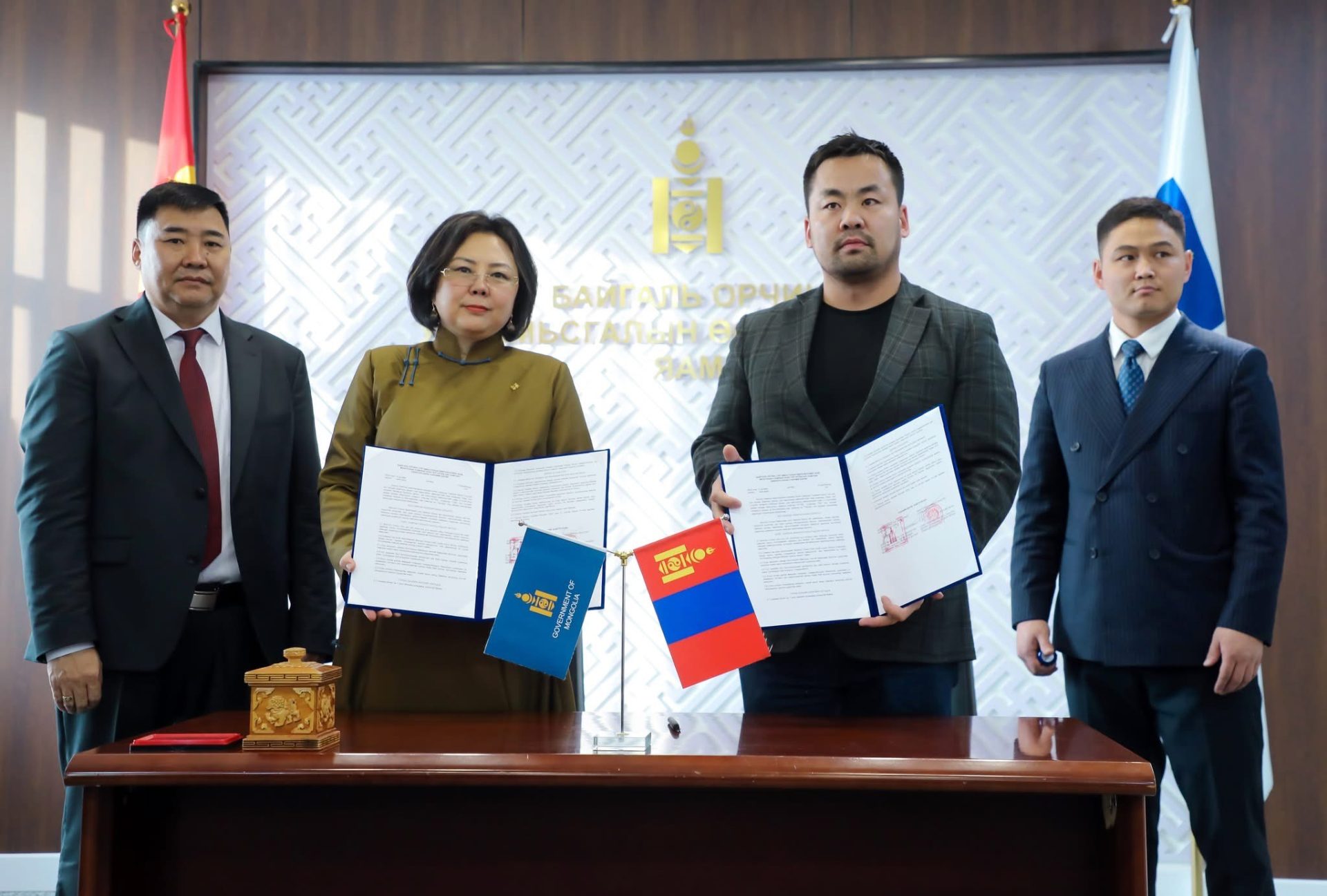 Four individuals stand around a table displaying documents and flags, including the Mongolian flag. They are in a formal setting with an emblem and intricate wall pattern in the background, signifying an official event or agreement.