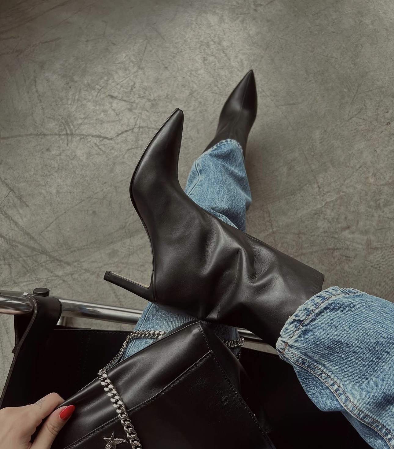 Person in light blue jeans wearing black pointed-toe high-heeled boots is sitting with legs crossed. The hand with red nails is holding a black leather bag with a metal chain on a gray surface.