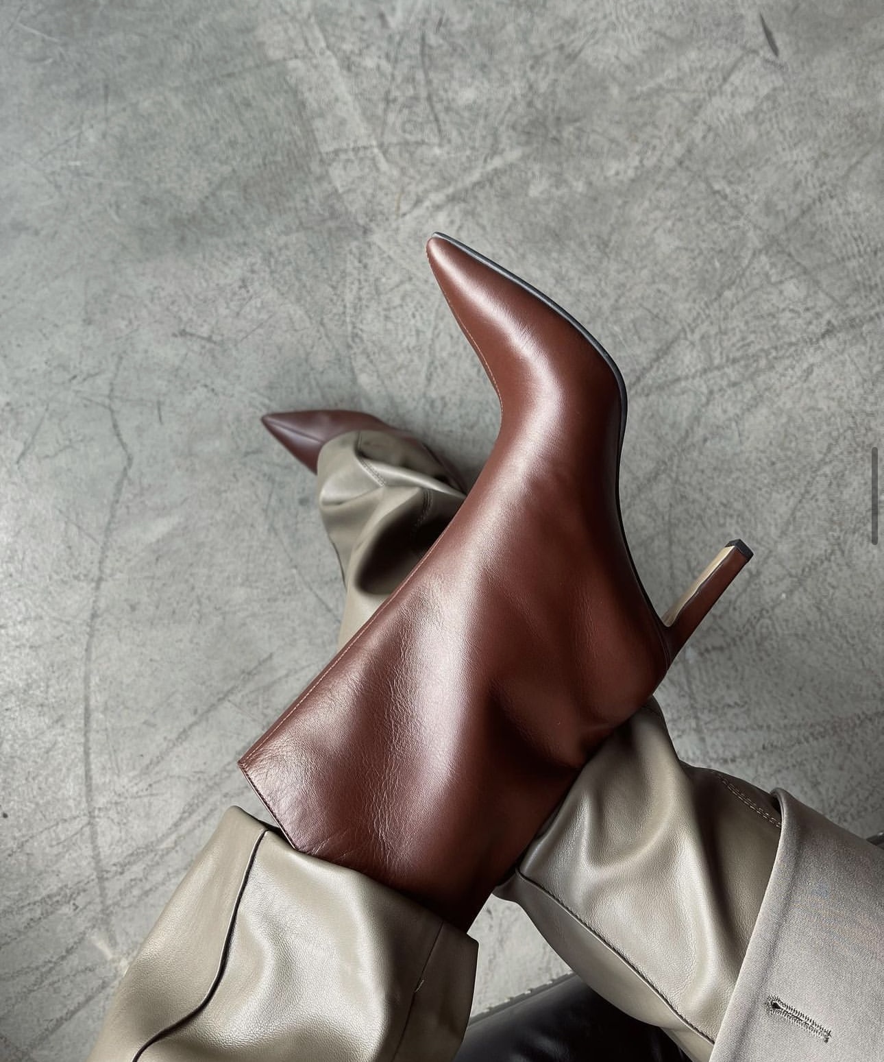 Close-up of a person wearing stylish brown leather pointed-toe boots with slender heels. The boots are paired with light gray pants, and the background features a textured gray surface.