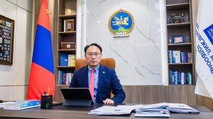 A person sits at a desk in an office with a tablet and books. The room features shelves with books and decorative items, a large flag, and an emblem on the wall. The individual wears a suit and glasses.