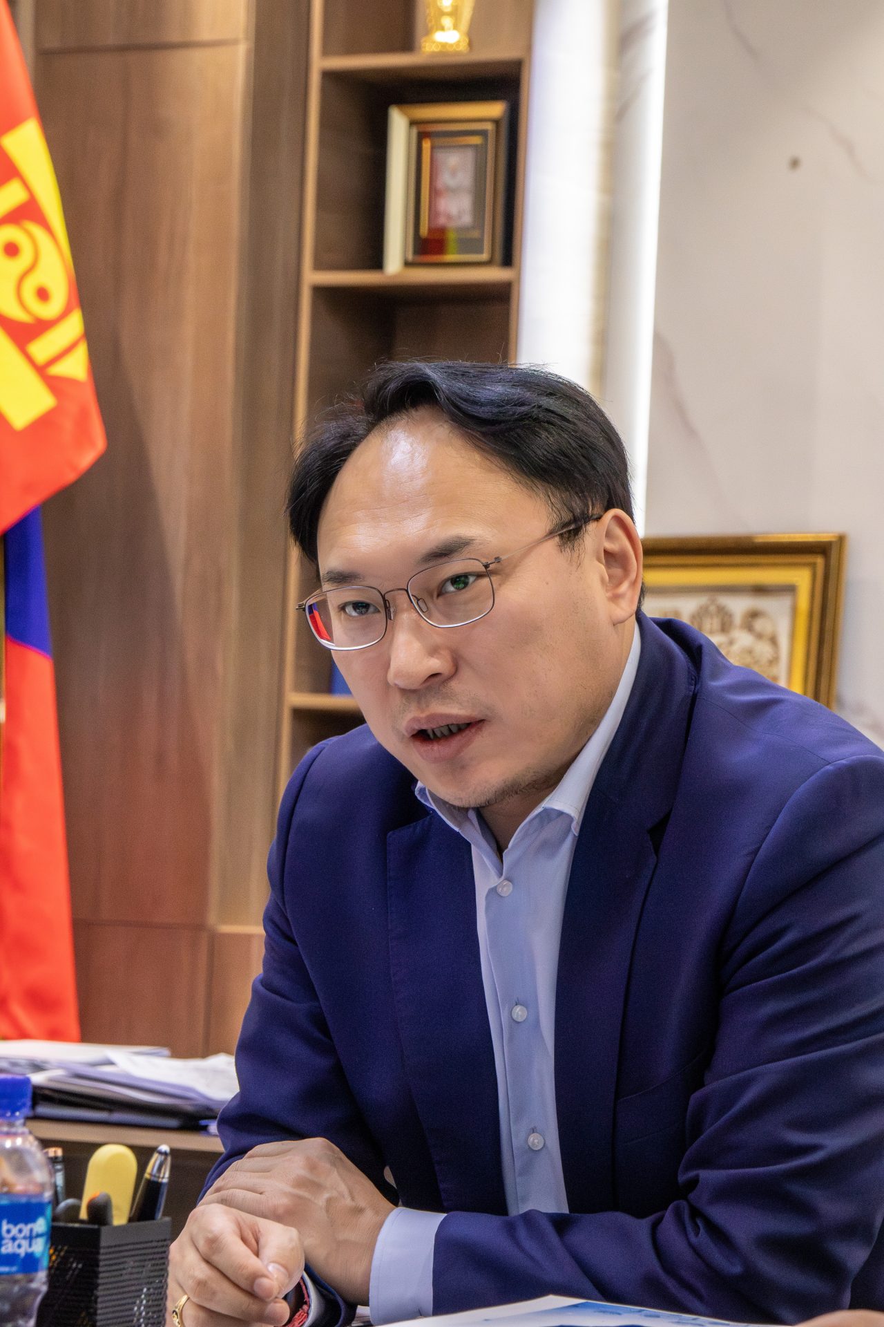 A man with glasses and a blue suit is seated at a desk in an office, speaking. A Mongolian flag and framed awards are visible in the background.