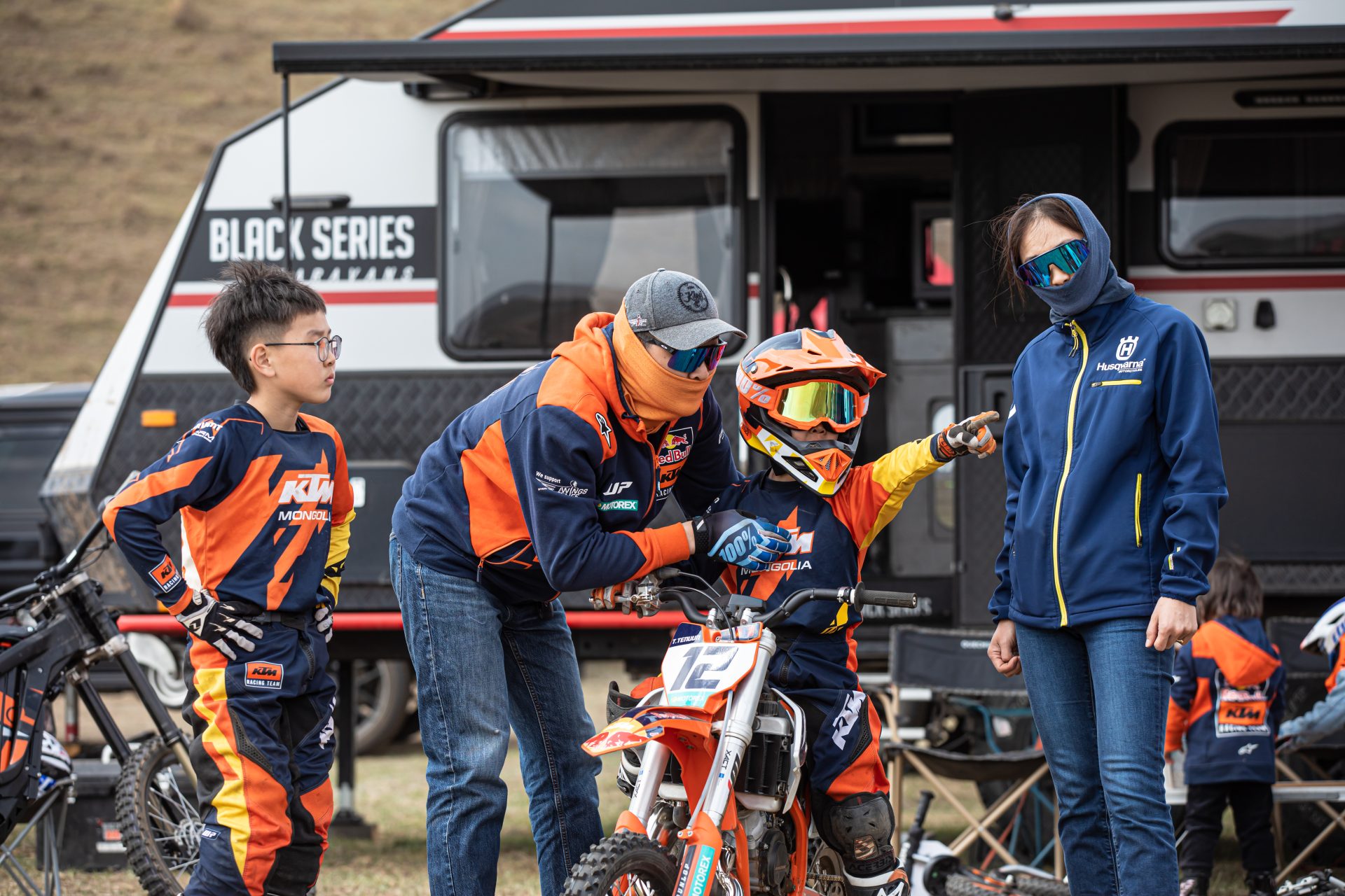 A child in motocross gear sits on a dirt bike, pointing ahead, surrounded by two adults and another child. A trailer with "Black Series" branding is in the background. The group is dressed in colorful racing outfits, preparing for motocross activity.