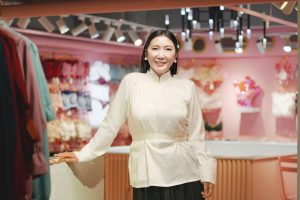 A woman with long dark hair stands in a brightly lit boutique. She's wearing a white blouse and black skirt, smiling at the camera. The shop is decorated in pink tones, with racks of clothing displayed in the background.