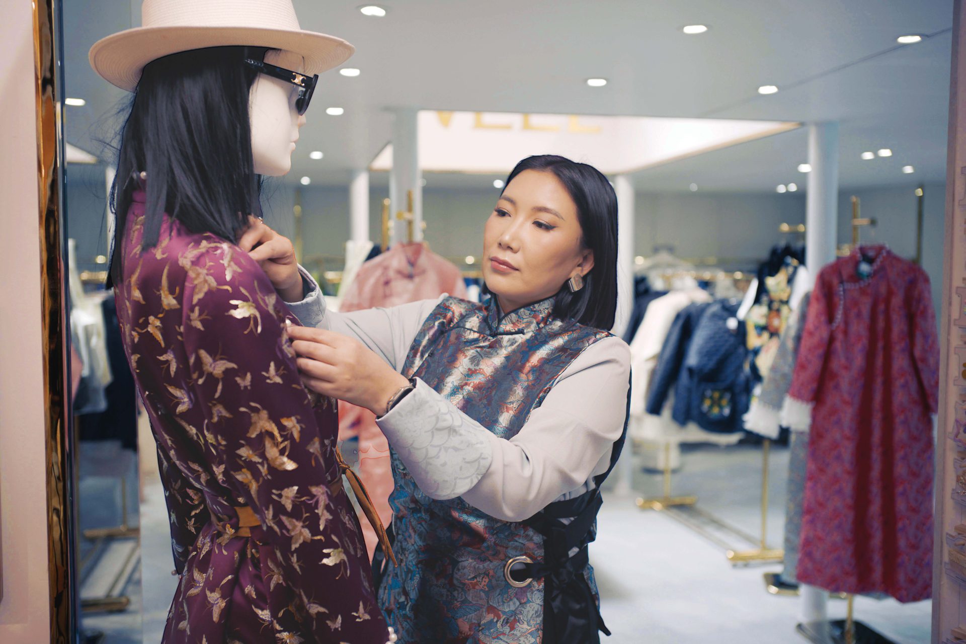 A woman adjusts the collar of a stylish dress on a mannequin in a clothing store. The store is filled with colorful garments on racks, and the woman is focused on perfecting the display.