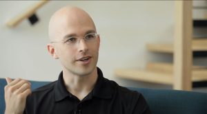 A person with glasses and a bald head is speaking and gesturing with their hand. They are wearing a black shirt and sitting on a blue sofa. The background shows a wooden staircase and light-colored walls.