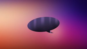 A dark speech bubble is centered against a gradient background transitioning from light pink and orange on the left to deep purple on the right. The bubble is empty, suggesting communication or conversation.