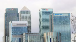 Skyline of modern skyscrapers with glass exteriors featuring logos from several financial institutions, including Citi, HSBC, Barclays, and State Street, under a cloudy sky.