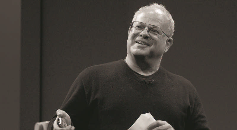 A man wearing glasses and a dark sweater is speaking and smiling, holding a piece of paper and remote. He's standing in front of a dark background, possibly giving a presentation.